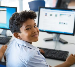 Young happy schoolboy using computer to search internet. Arab child learning to use computer at elementary school. Portrait of smiling middle eastern kid looking at camera while surfing the net in school library.