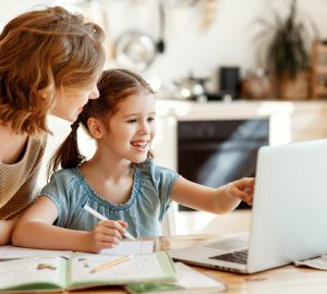 Positive young woman helping daughter in searching information for homework on internet while sitting together at table with laptop at home