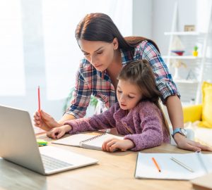 Mother and daughter looking at notebook near laptop on desk at home on blurred background