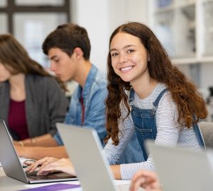 Happy young woman working on laptop and looking at camera in classroom. Portrait of smiling university student in library use computer for a research. Satisfied college student looking at camera while sitting in a row with classmates studying together.