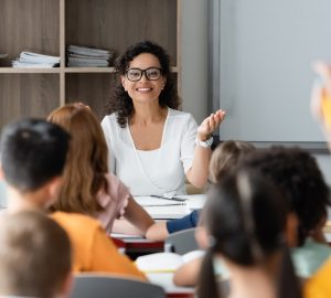 positive african american teacher pointing at blurred girl raising hand in classroom